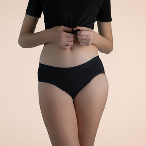 A teen girl wearing black Sahara period proof underwear boyleg style. Heavy absorbency. Model is wearing a black top. Off-white background. Size Y13-14 years. Front angle.