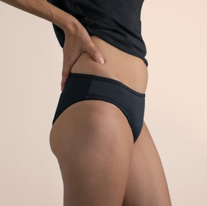 A female wearing black Sahara period proof swimwear bikini brief style. Model is wearing a black top. Off-white background. Size 8/XS. Side angle.