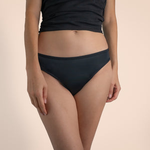 A female wearing black Sahara period proof swimwear bikini brief style. Model is wearing a black top. Off-white background. Size 8/XS. Front angle.