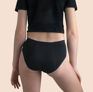 A teen girl wearing black Sahara period proof underwear boyleg style. Heavy absorbency. Model is wearing a black top. Off-white background. Size Y12-13 years. Back angle.