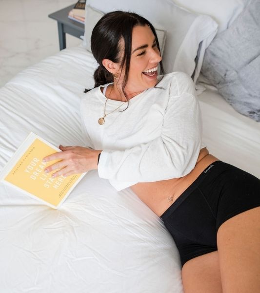 Woman lying on a bed holding a book and laughing. Wearing a white sweatshirt and Sahara period black shorts.
