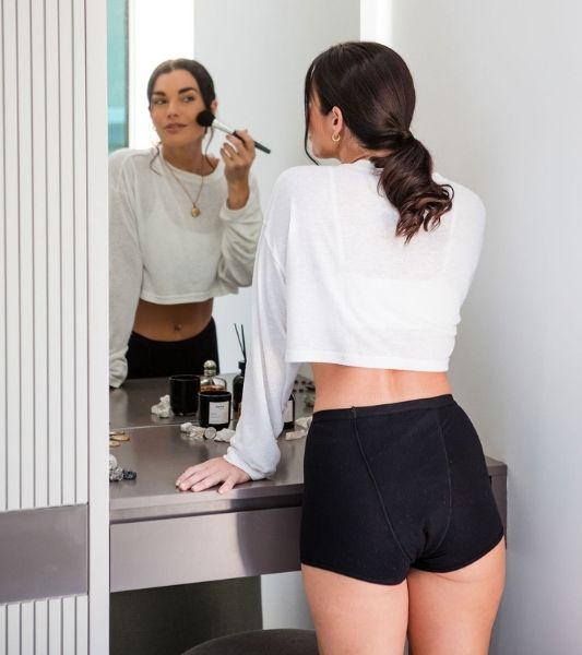 Woman applying makeup in front of a mirror, wearing a white top and Sahara period pants in boyshort style.