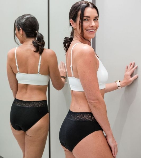 Woman wearing a white bra and black Sahara lace period pants, standing against a plain background.